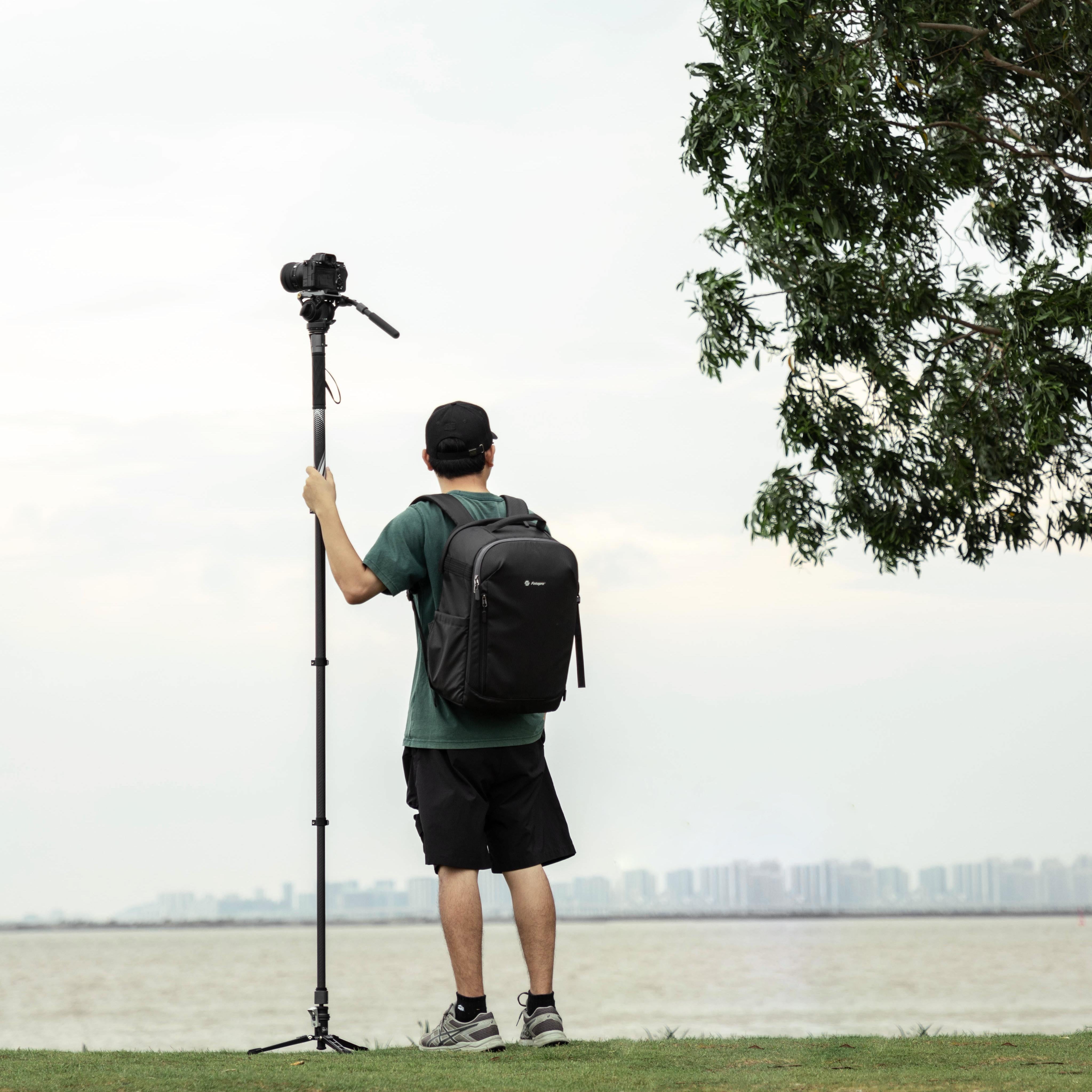 Person with camera mounted on fotopro x-airfly mono 3-in-1 monopod and video head and backpack standing outdoors near the water.